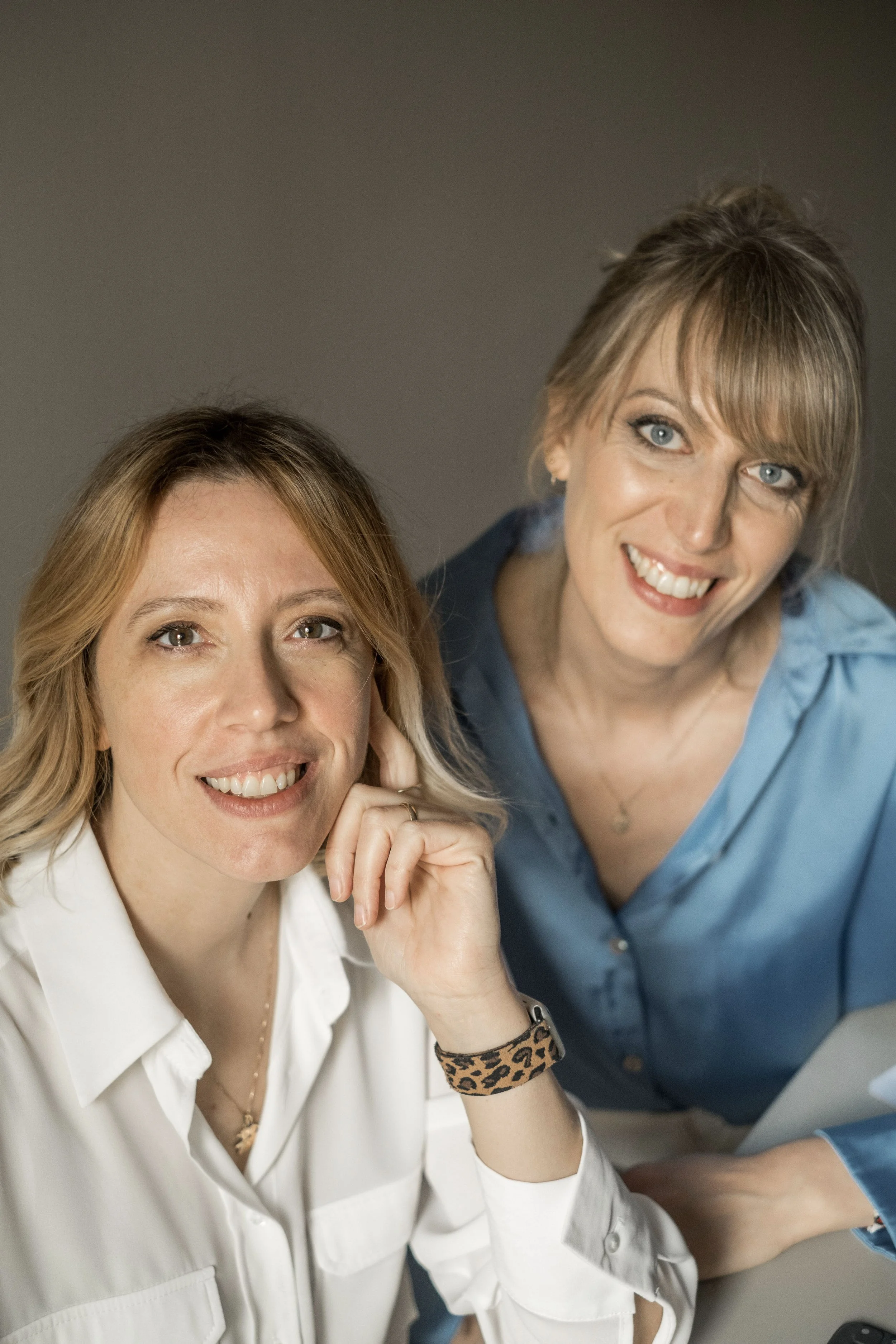 Due donne sorridenti in primo piano, una con capelli castani chiari e l'altra con capelli castani scuri, indossano camicie casual e sono in un ambiente neutro.