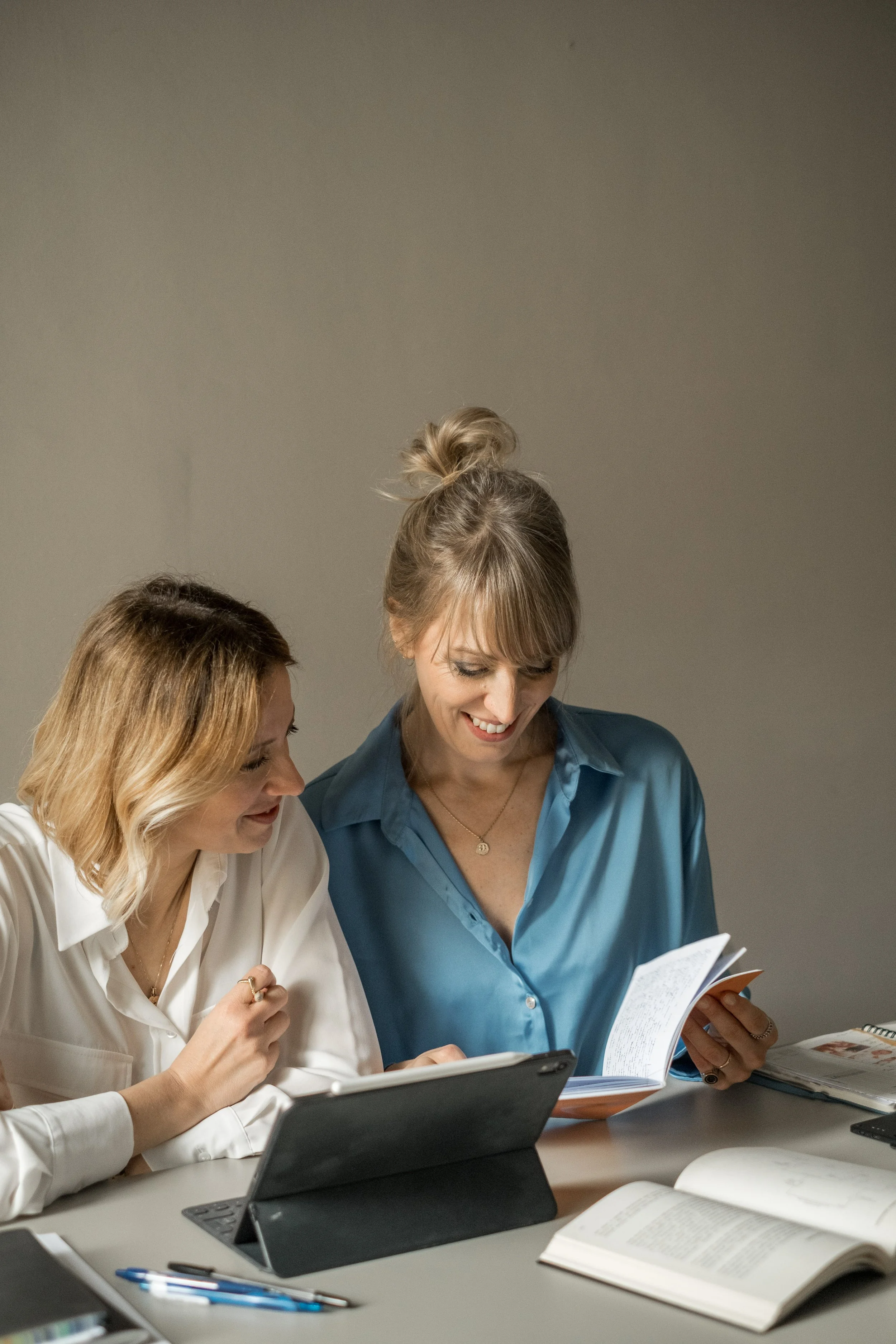 Due donne sedute a un tavolo, una con una copia di un libro e l'altra con un tablet, che discutono e sorridono durante uno studio o una riunione.