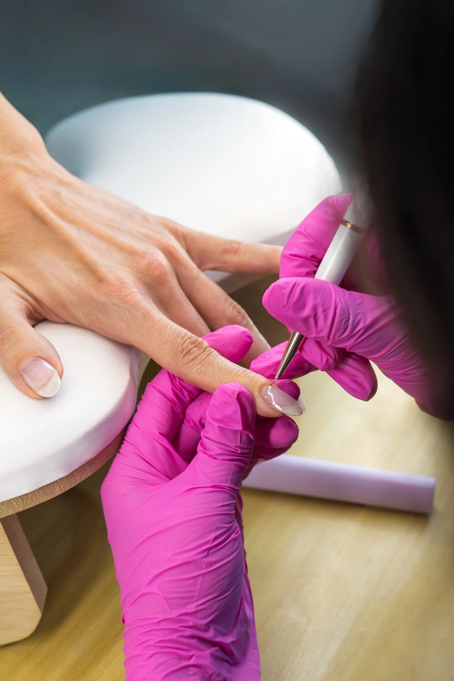 Close-up of a person receiving a professional manicure, with the technician applying nail art using a pen tool. The person's hand is resting on a white cushion, and the technician is wearing bright pink gloves.