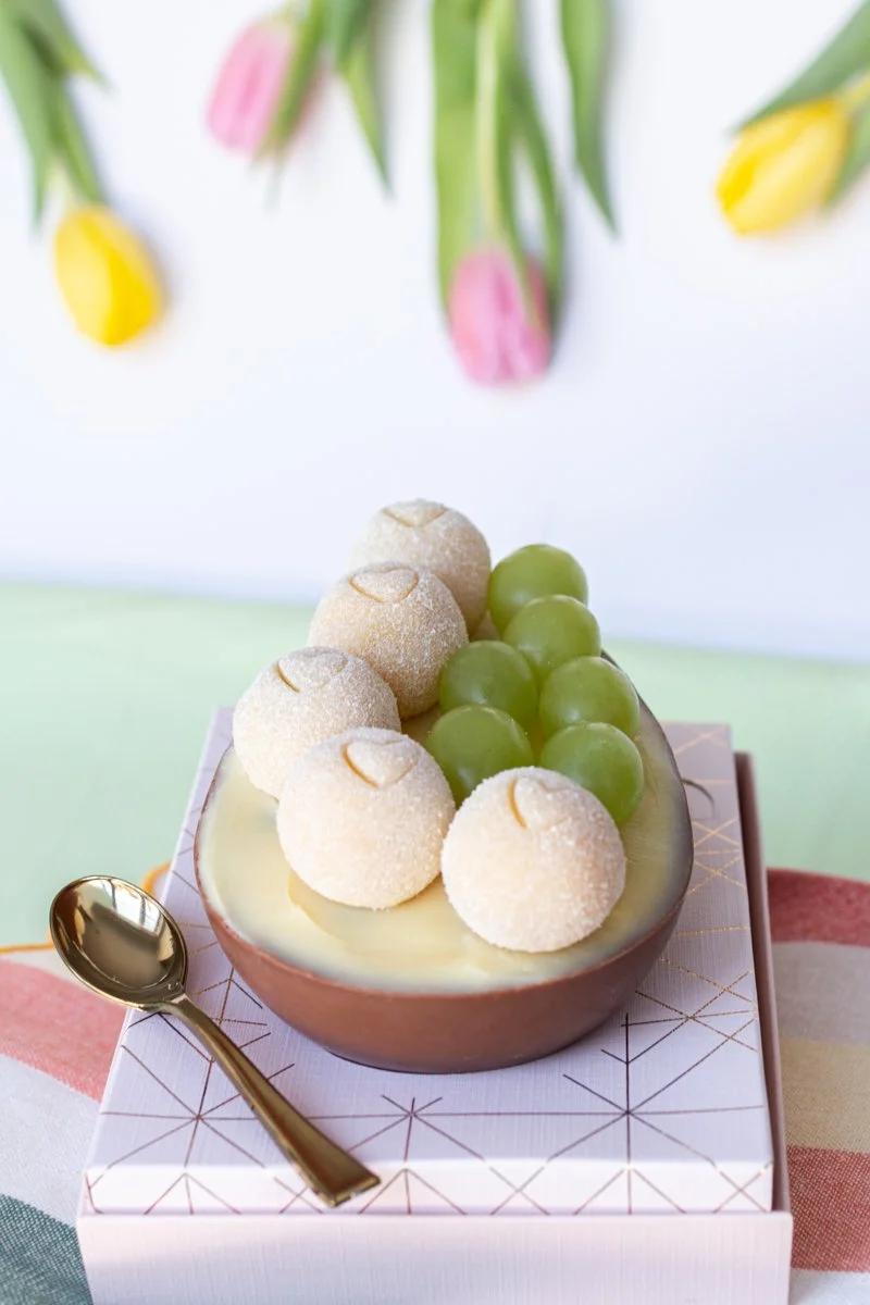 Dessert bowl with white round truffles, green grapes, and white chocolate base, placed on a geometric-patterned box, with a gold spoon beside it, and colorful tulips hanging in the background.