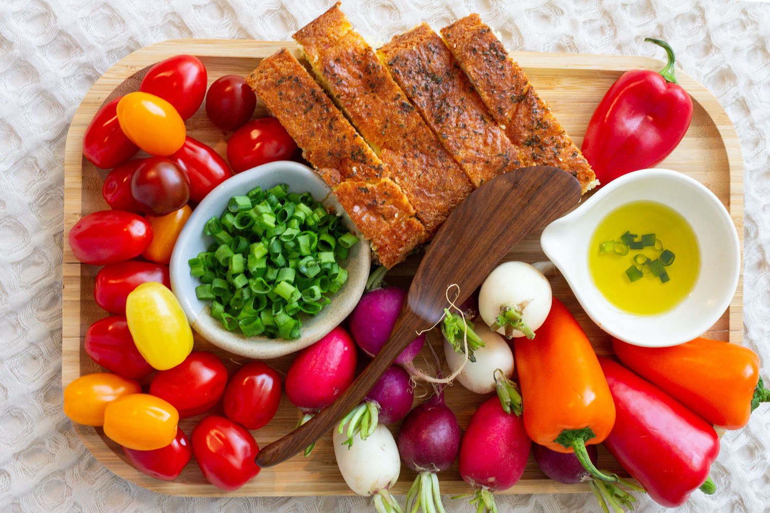 A wooden tray with grilled fish fillet, cherry and heirloom tomatoes, colorful radishes, green onions, a small bowl of chopped green onions, a bowl of olive oil with chopped green onions, and assorted bell peppers in red, orange, and yellow.