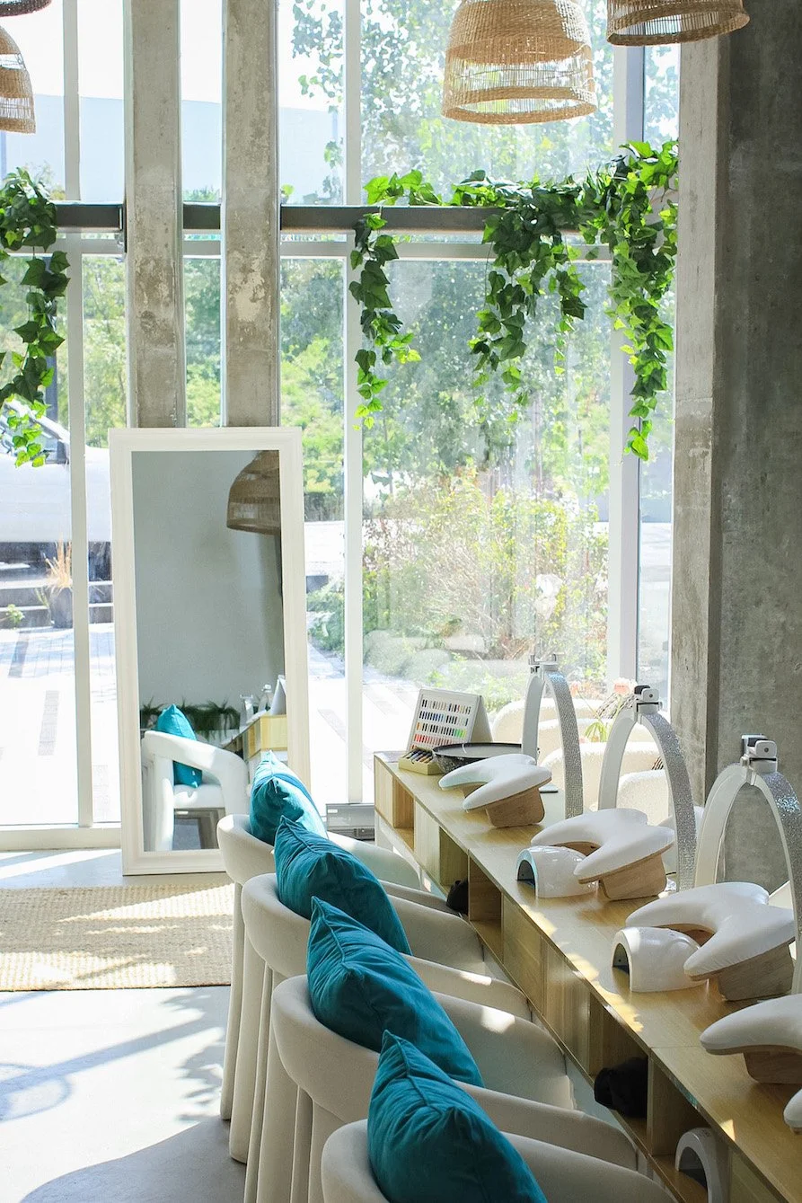 Interior of a manicure or pedicure salon with massage chairs, teal pillows, and nail polish devices, illuminated by natural light through large windows decorated with hanging plants.