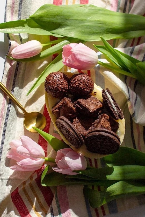A white bowl filled with assorted chocolates, including Oreo cookies and chocolate truffles, on a striped tablecloth. Pink tulips and green leaves are arranged around the bowl, with a gold spoon placed on the left side.