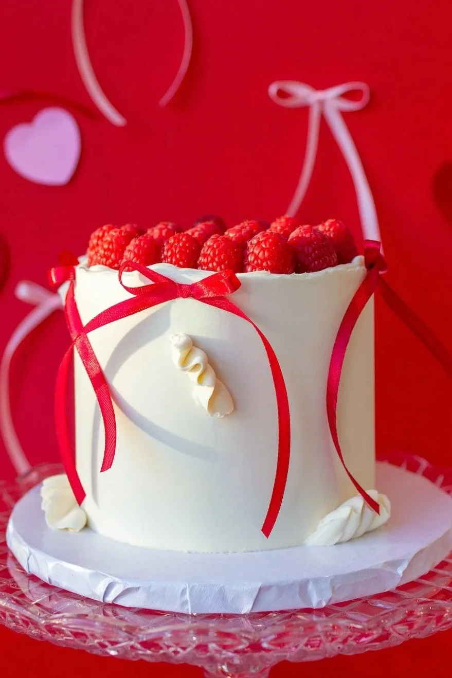 A white birthday cake decorated with raspberries on top and red ribbons on the sides, on a glass cake stand with a red background featuring white and pink hearts and ribbons.