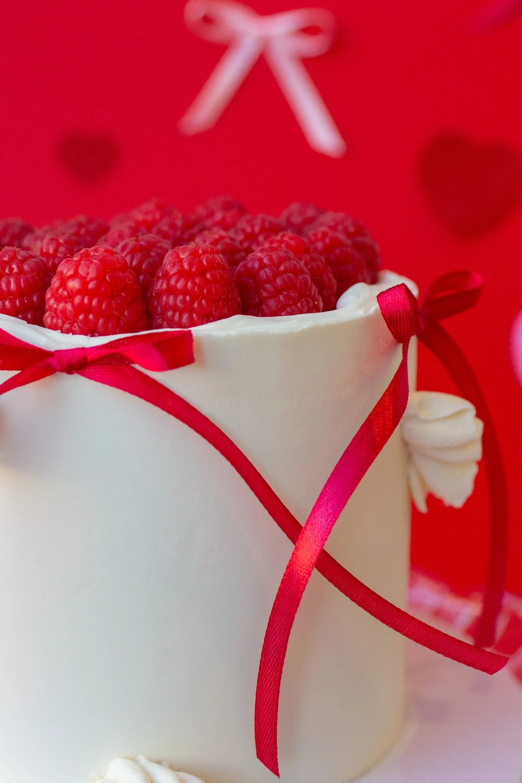 A white cake decorated with red berries on top and red ribbons on the sides, with blurred red background