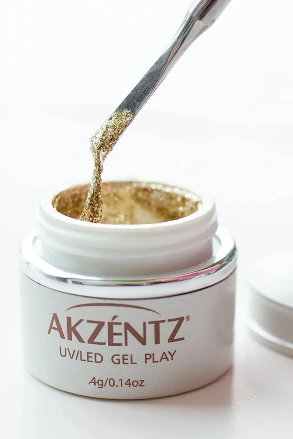 A white jar of Akzéntz UV/LED gel play with gold glitter gel inside and a brush picking up the gel.