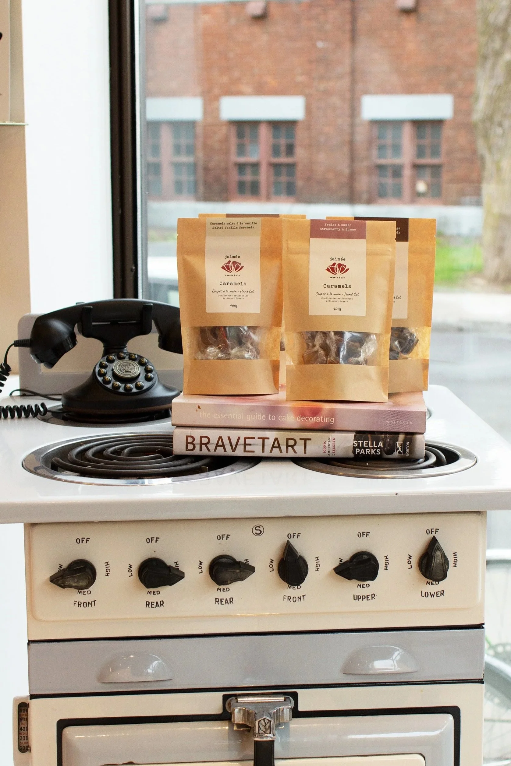 Vintage white stove with four knobs, three small books stacked on top, including 'Bravetart', 'the essential guide to cake decorating', and 'Stella Parks'. Three bags of caramel candies on top of the books. A black rotary phone is on the left side of