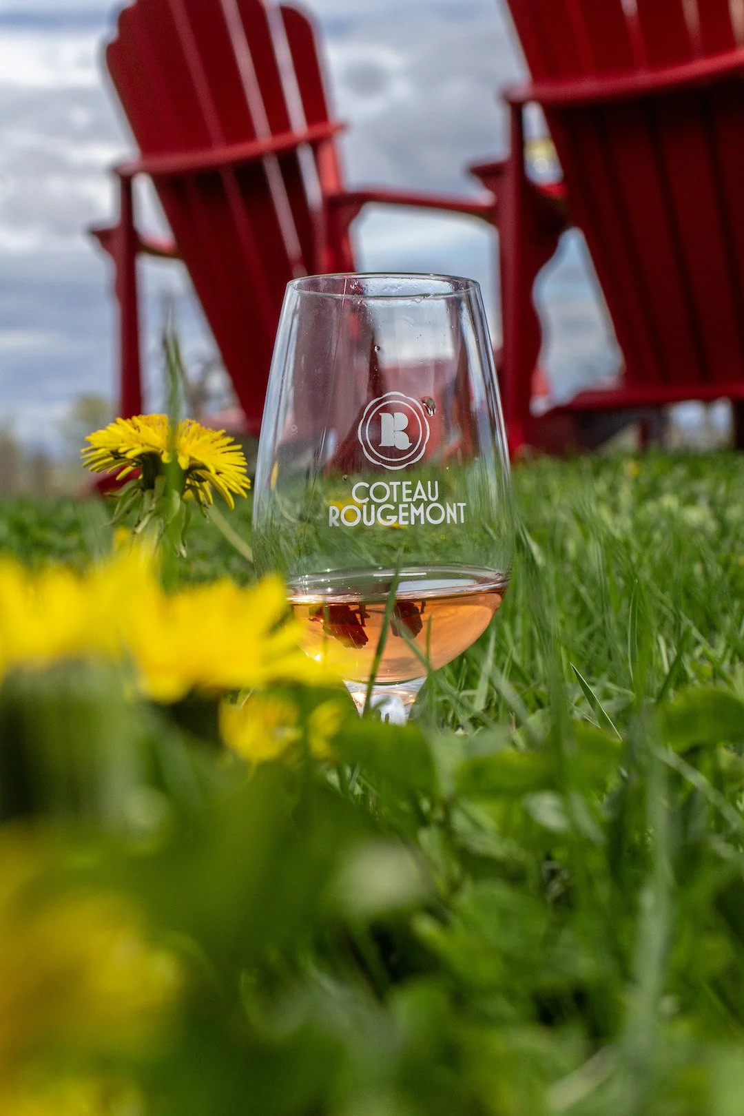 A glass of pink rosé wine with the label 'Coteau Rougemont' on the grass, with yellow flowers in the foreground and red Adirondack chairs in the background, under a partly cloudy sky.