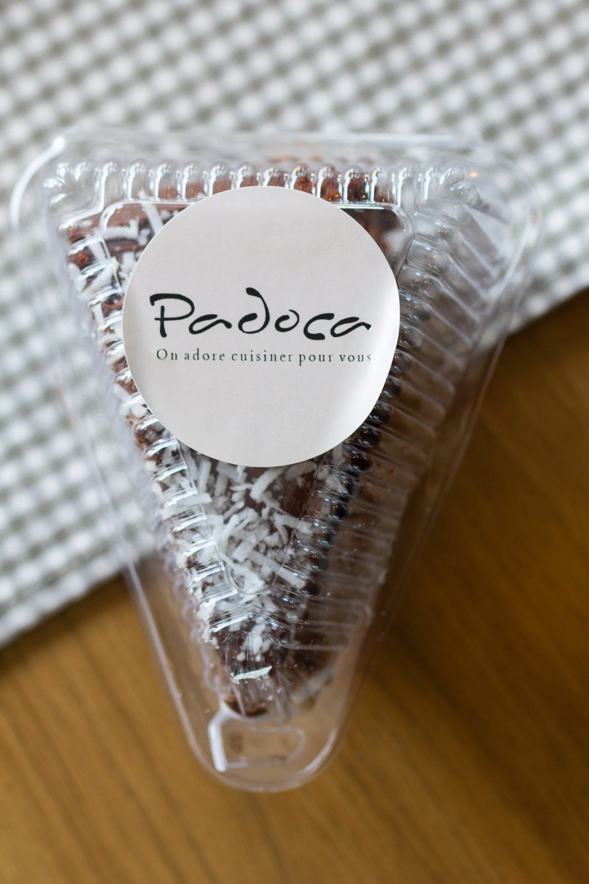 Clear plastic container with chocolate cake, topped with shredded coconut, labeled 'Paddoca' with a French phrase 'On adore cuisiner pour vous'.
