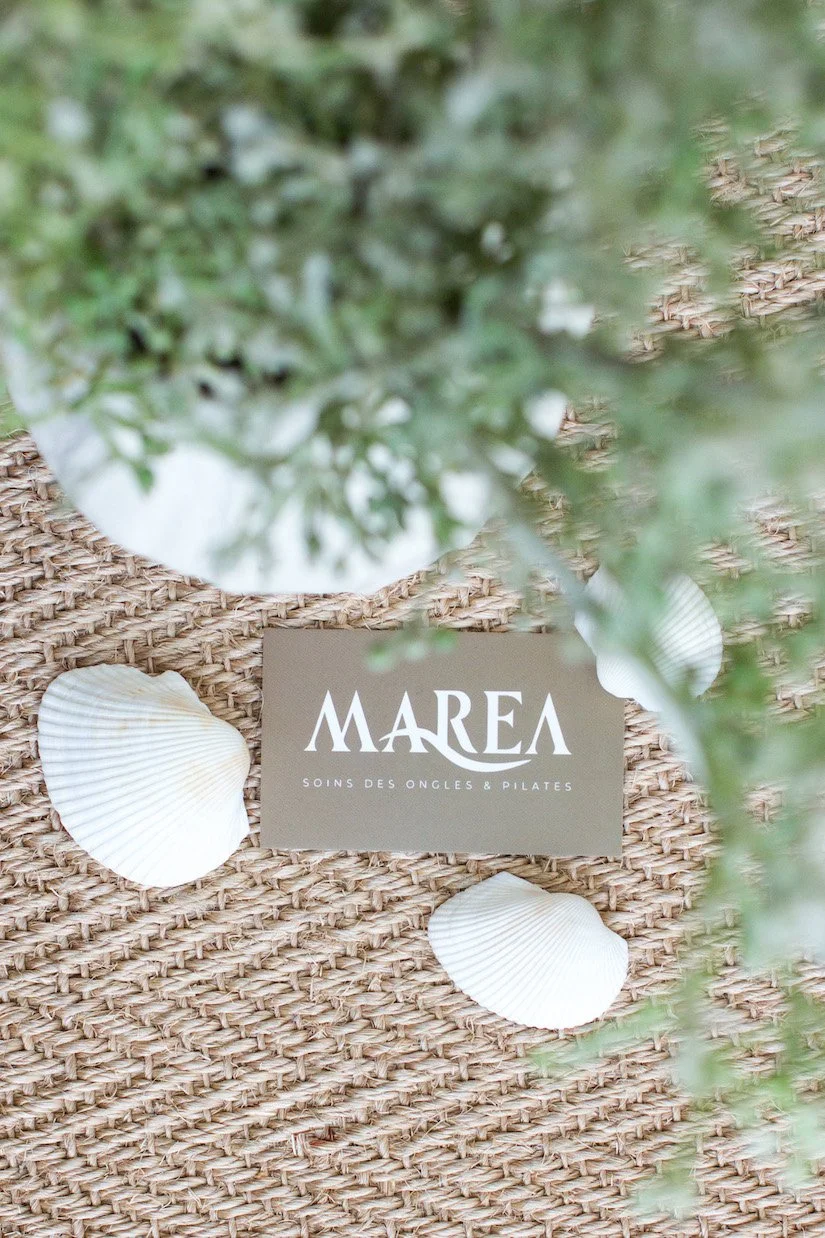 A business card for MAREA, a nail and Pilates studio, placed on woven surface with seashells surrounding it and greenery partially obscuring the view.