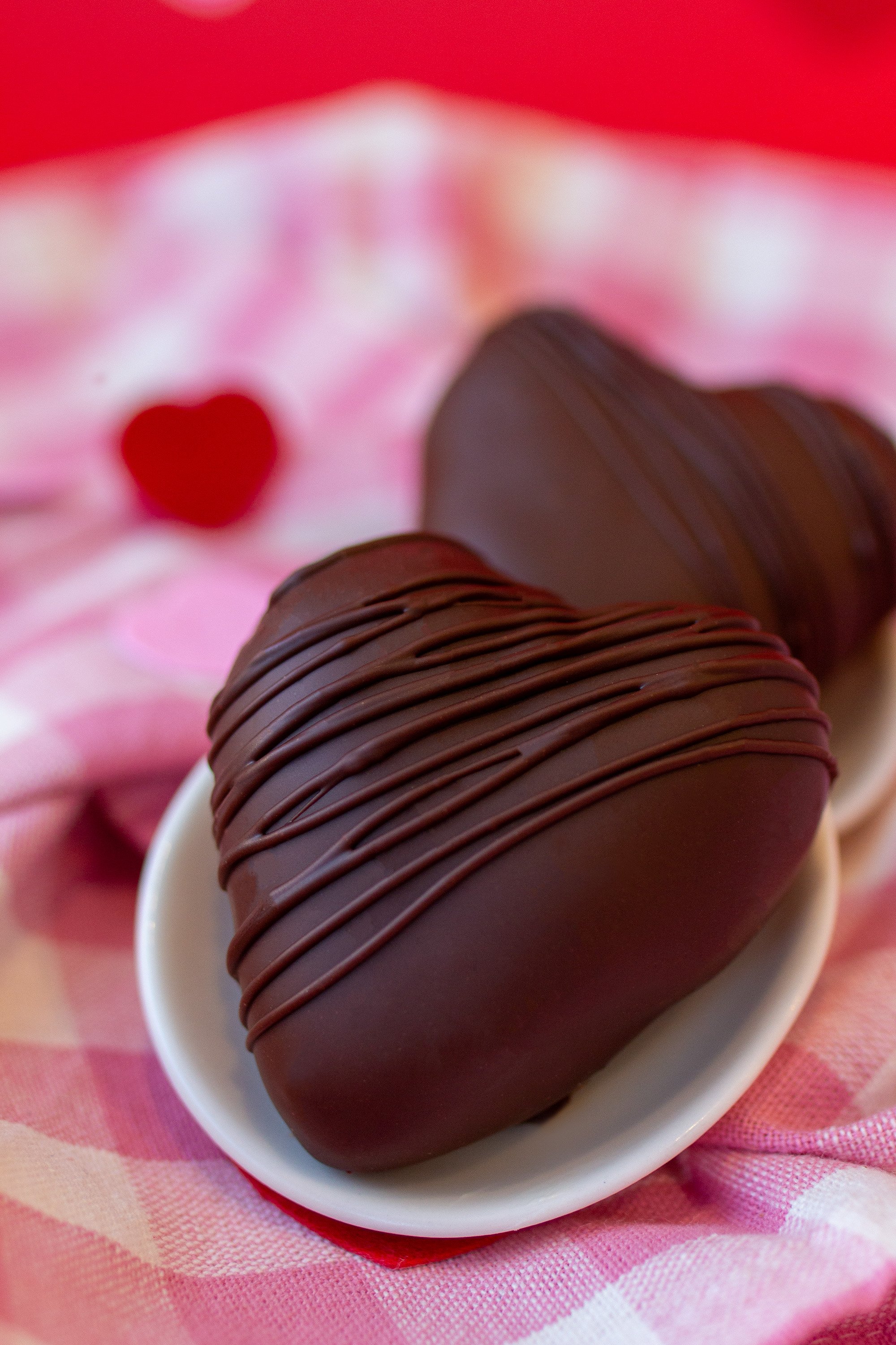 Two heart-shaped chocolates coated in dark chocolate with thin chocolate drizzle, placed on a small white dish, background with a pink and red theme and a blurred red heart.