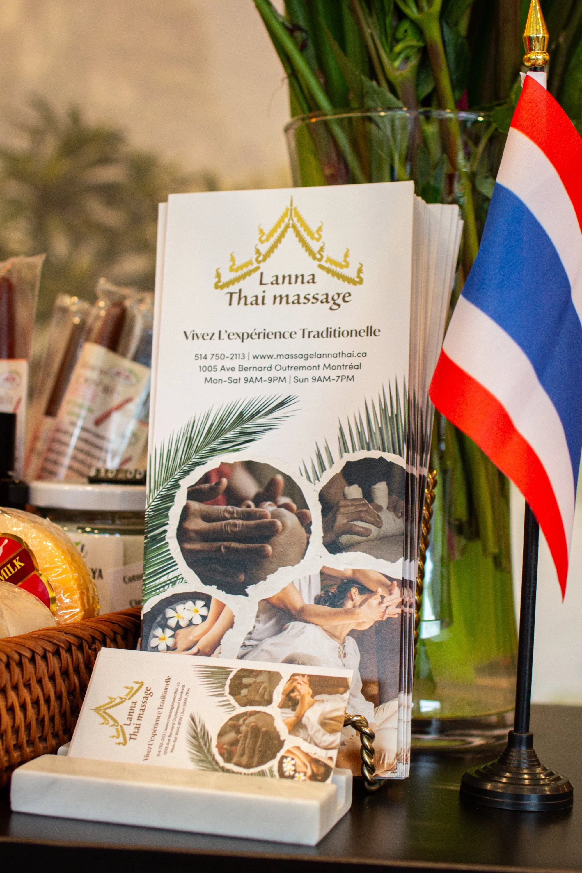 Brochure for Lanna Thai massage with images of Thai massage techniques, a Thai flag, and contact information, placed on a table next to a basket of snacks.