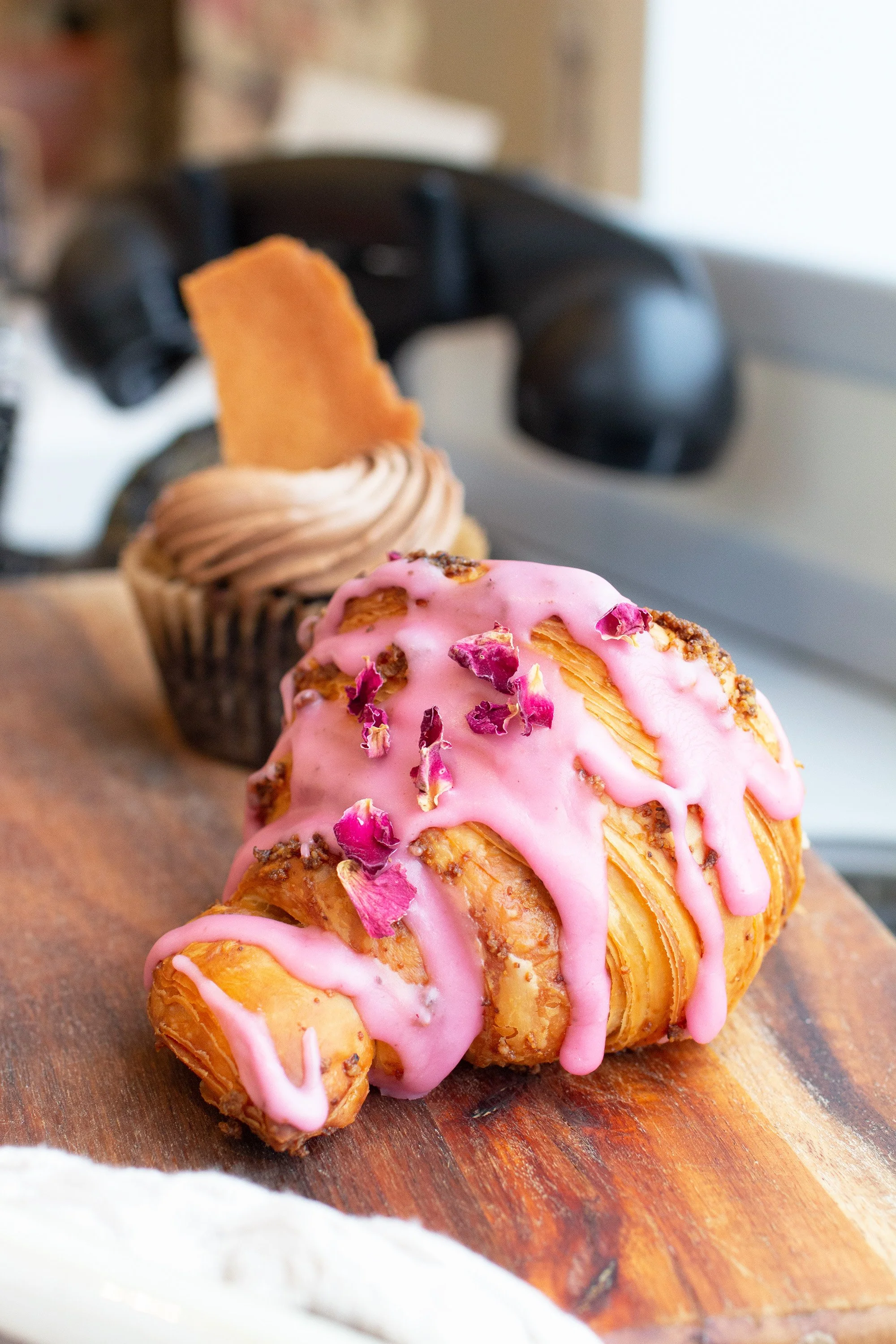A pink glazed croissant with dried rose petals on top, placed on a wooden surface with a cupcake topped with a swirl of frosting and a cookie in the background.