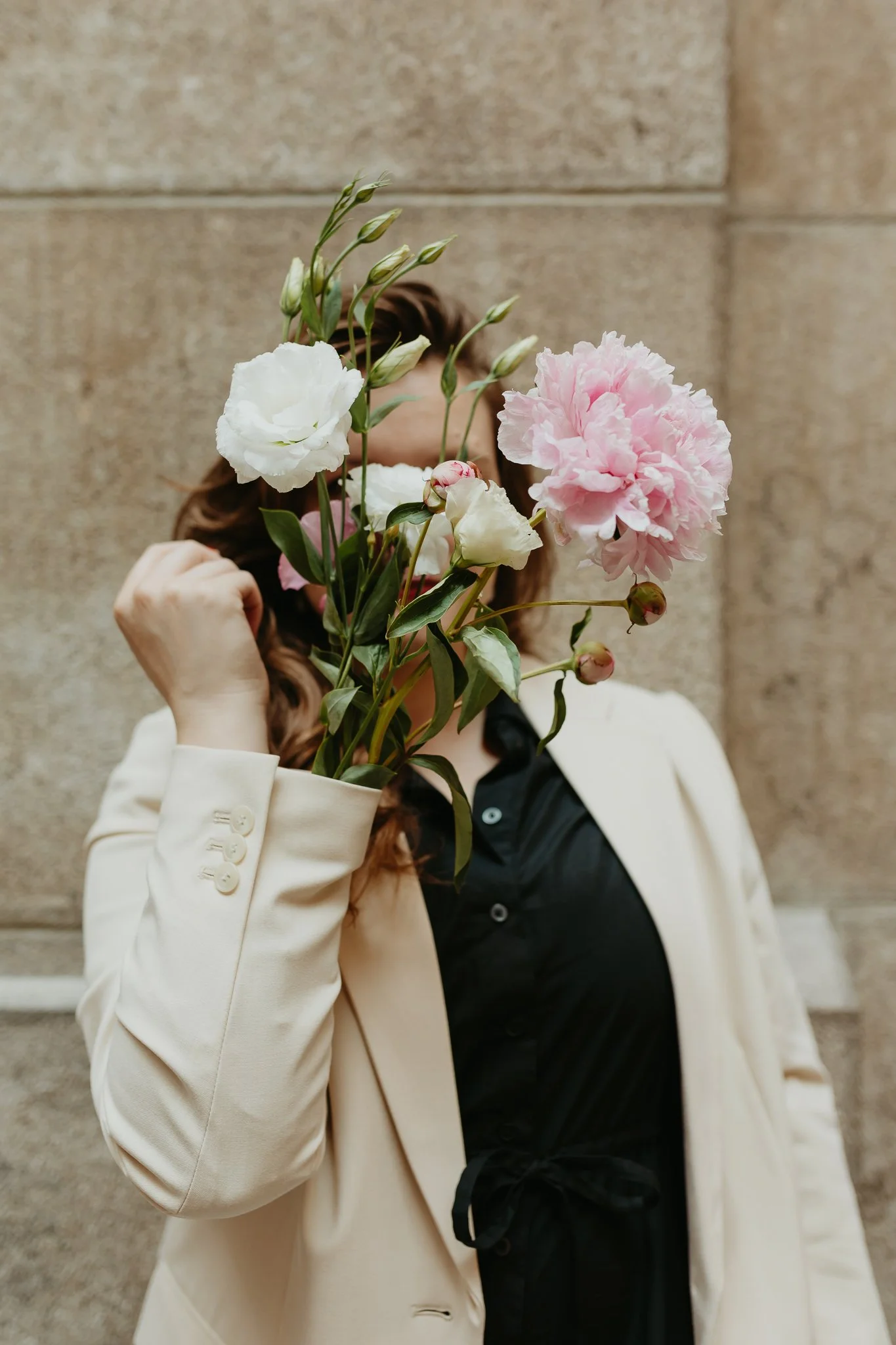 A woman holding a bouquet of pink and white flowers in front of her face, wearing a cream blazer and black shirt, standing in front of a stone wall.