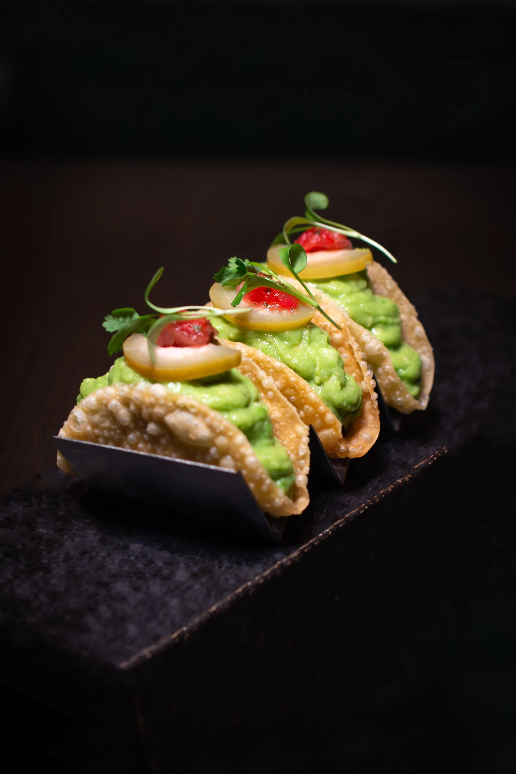 Three gourmet tacos with green filling, topped with sliced vegetables and microgreens, arranged on a black slate serving tray.