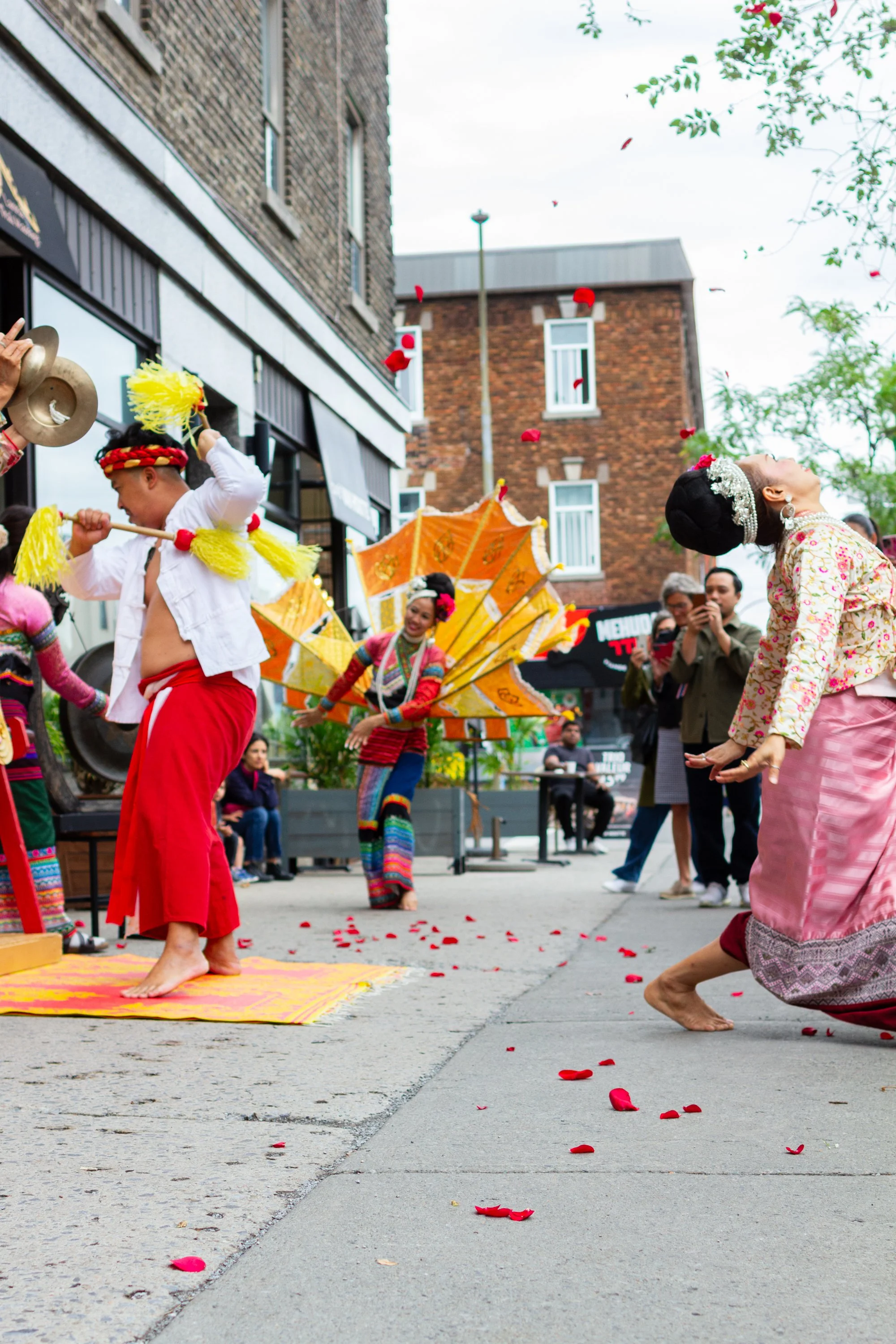 People in traditional Asian costumes dancing and playing instruments during a street performance, with flower petals scattered on the ground and onlookers watching.