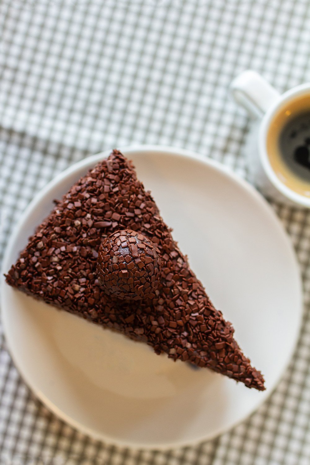 A slice of chocolate cake topped with chocolate shavings on a white plate, with a cup of coffee in the background.