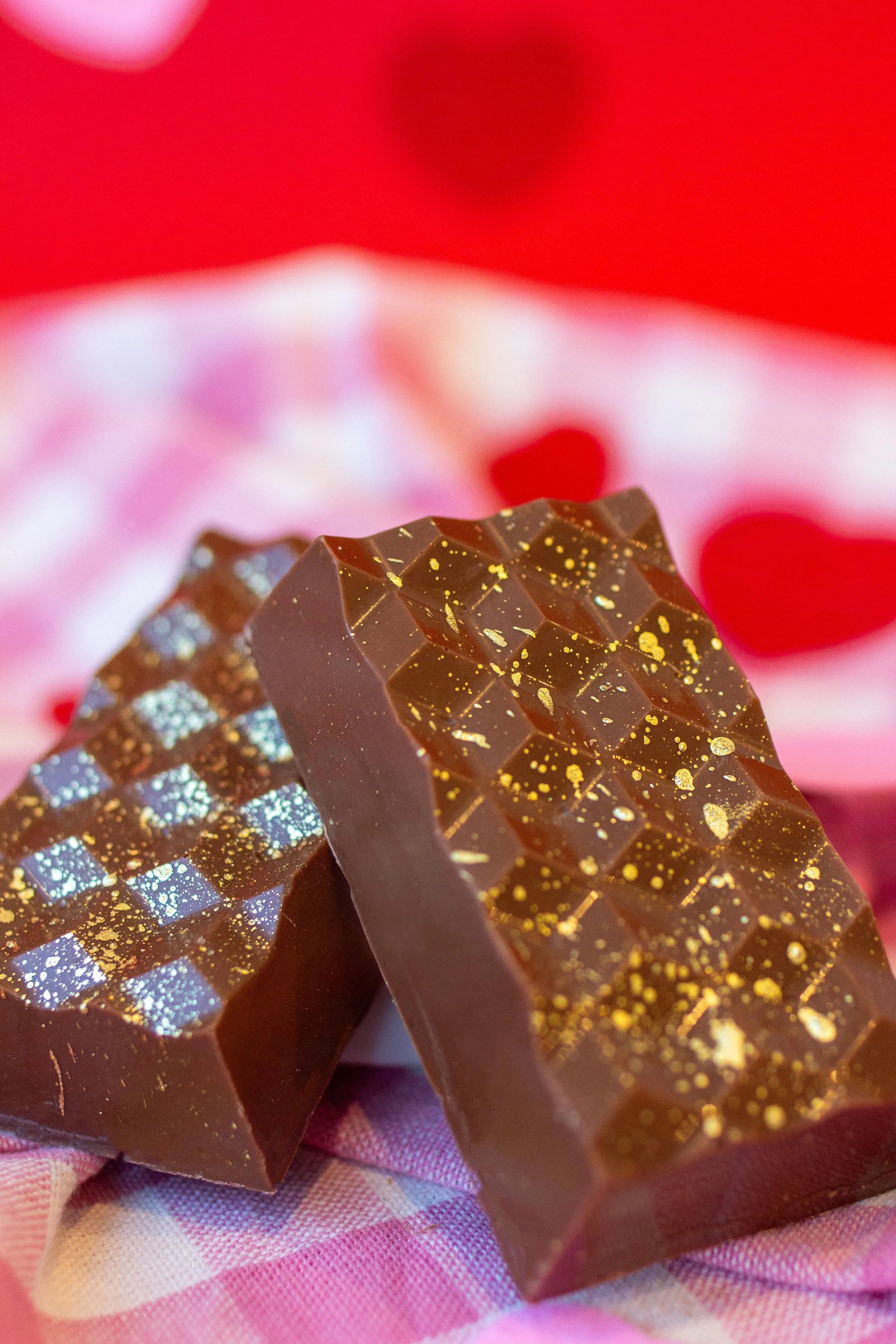 Close-up of two pieces of dark chocolate with gold leaf and glitter decorations, placed on a pink and white checkered cloth.