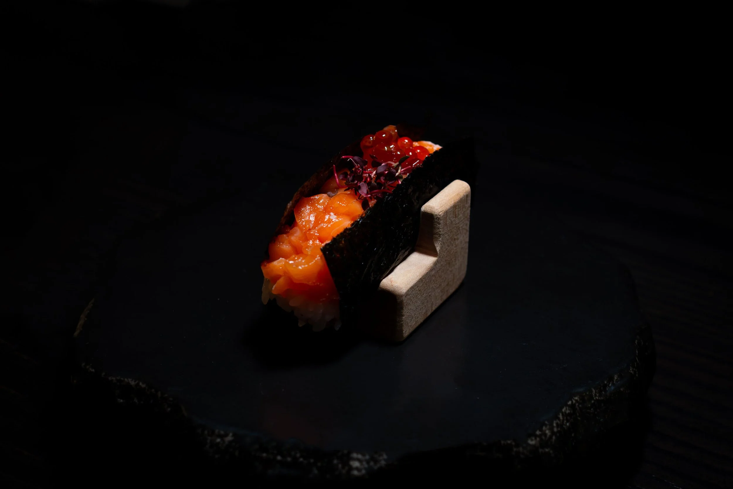 A piece of salmon sashimi wrapped in seaweed, garnished with salmon roe and microgreens, served on a small wooden stand on a black slate surface.