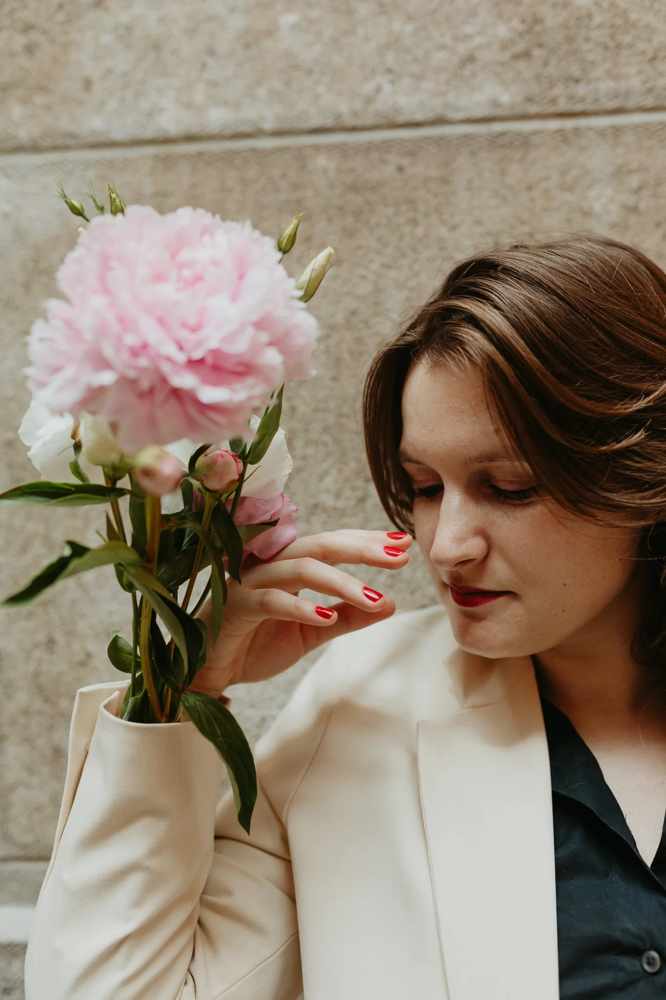 A woman with short brown hair, wearing a cream blazer and dark top, holding a pink and white flower close to her face, against a brick wall background.