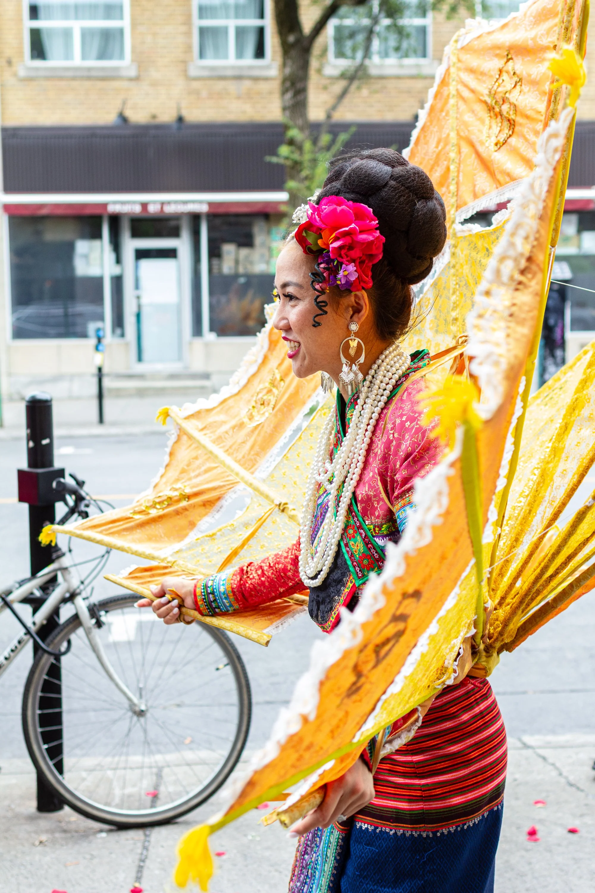 A woman dressed in traditional Mexican attire, holding a large bright yellow and orange parasol, standing on a city street with a bicycle nearby.