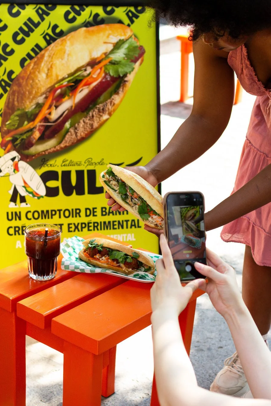 Person taking a photo of a banh mi sandwich on an orange table, with a drink and another sandwich, in front of a bright yellow sign advertising banh mi, outdoors on a sunny day.