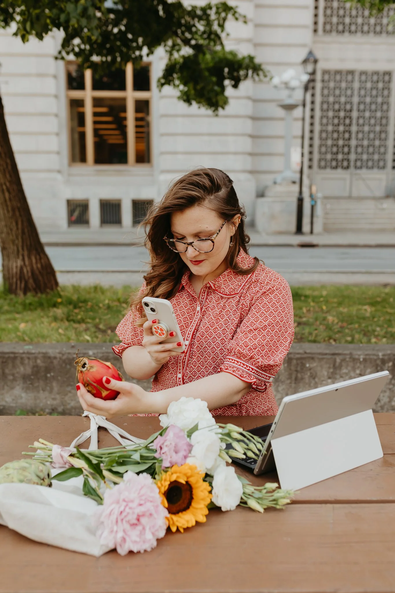 A woman in red patterned shirt with glasses, sitting outdoors at a wooden table, holding a pomegranate in one hand and looking at her phone with the other, with flowers and a tablet on the table.