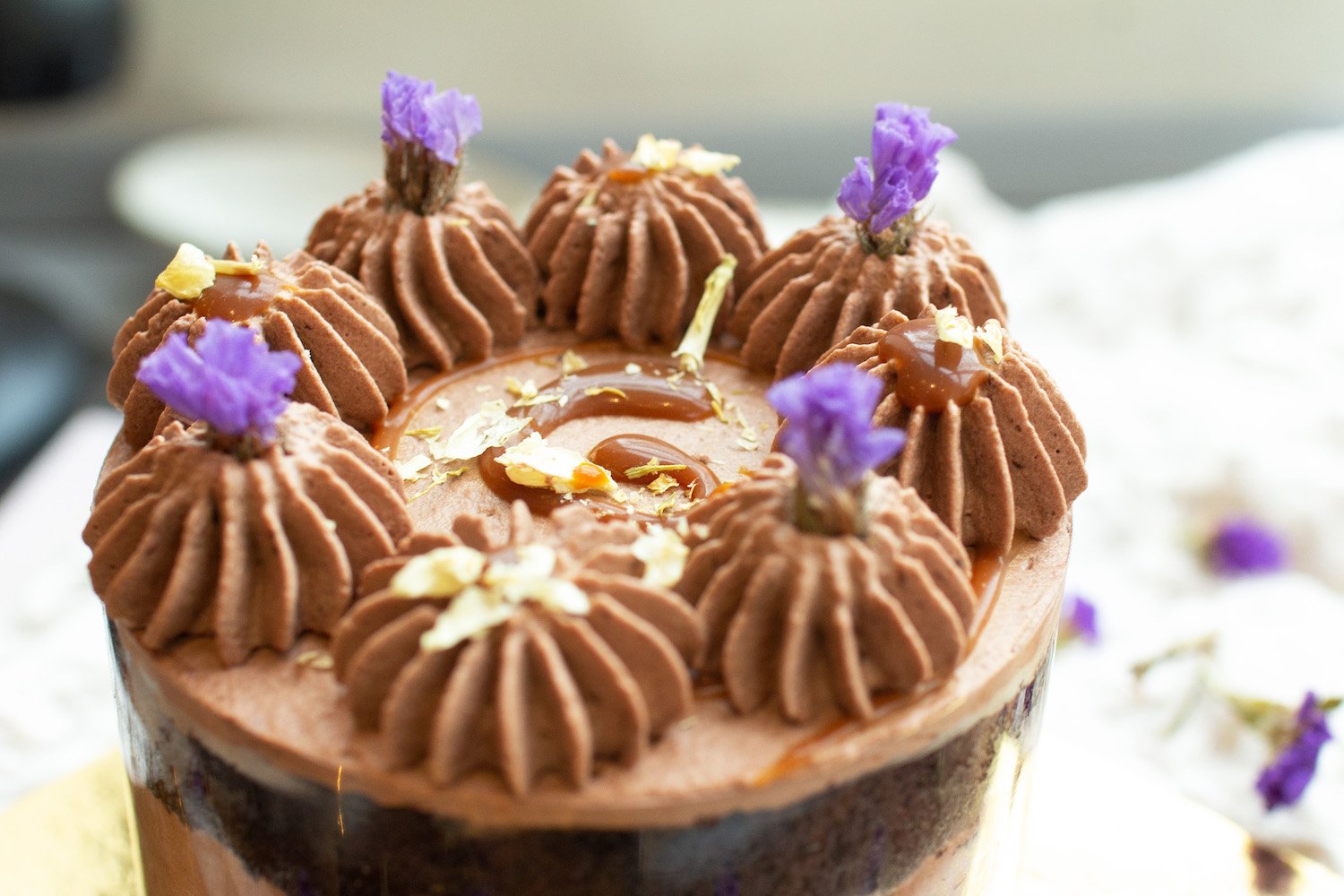 A chocolate cake decorated with piped chocolate frosting, edible purple flowers, caramel sauce, and white chocolate shavings.