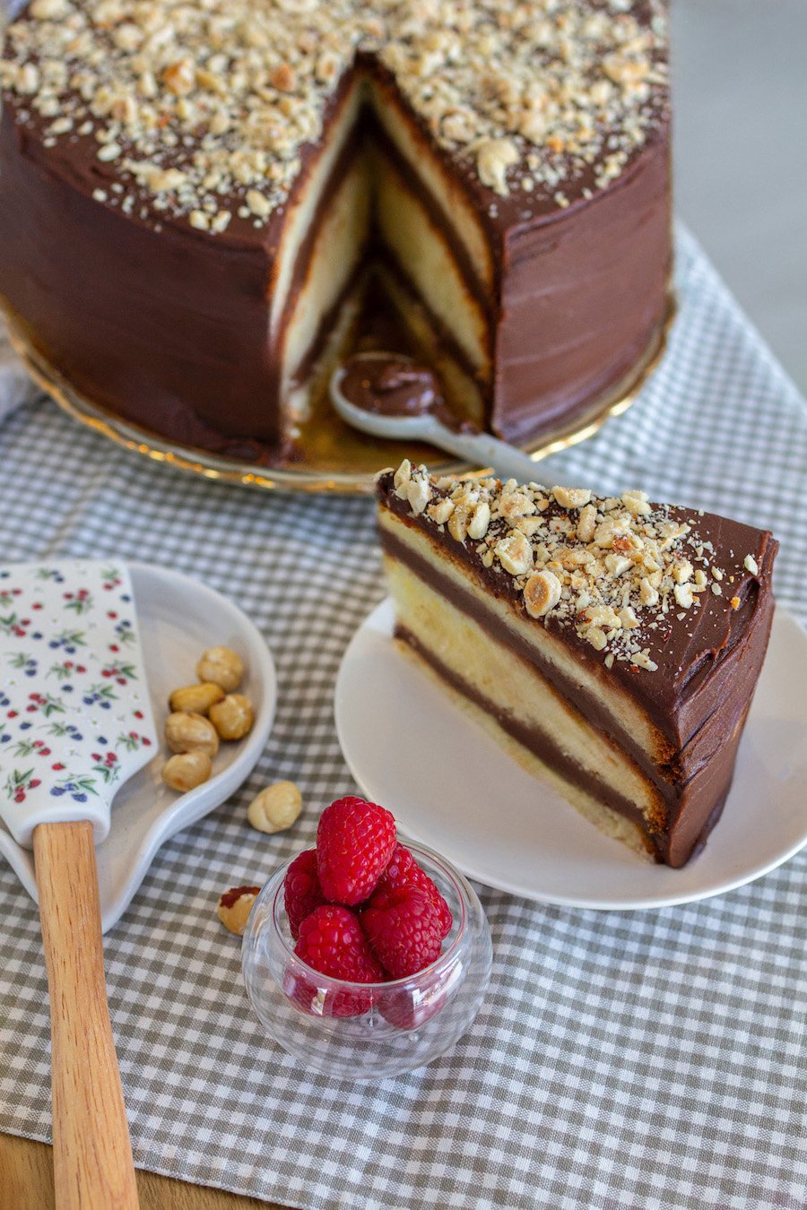 Slice of layered chocolate and vanilla cake with chopped nuts on top, on white plate, with raspberries in a glass, hazelnuts, and a spatula with floral design on a checkered tablecloth.