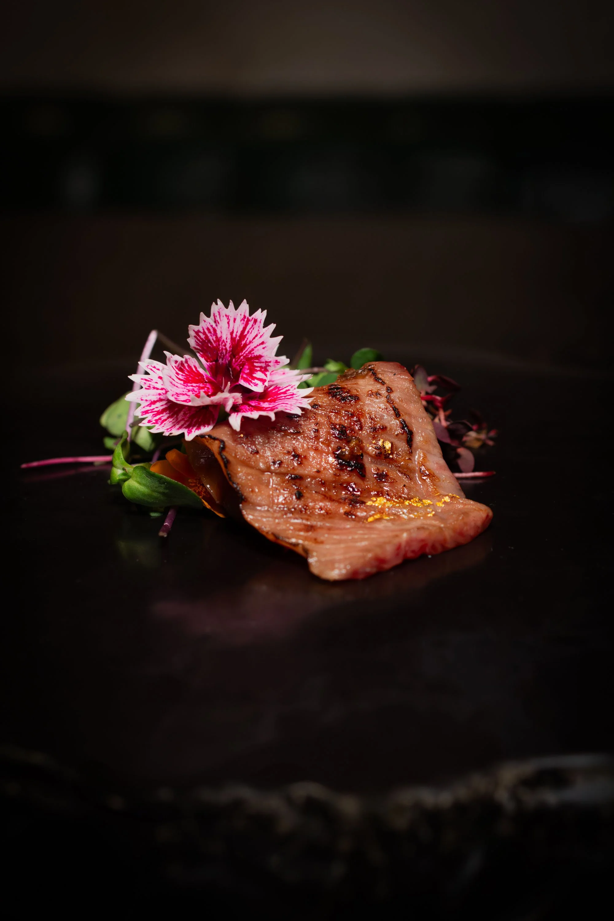 A grilled piece of meat garnished with pink and white edible flowers and microgreens, on a dark background.