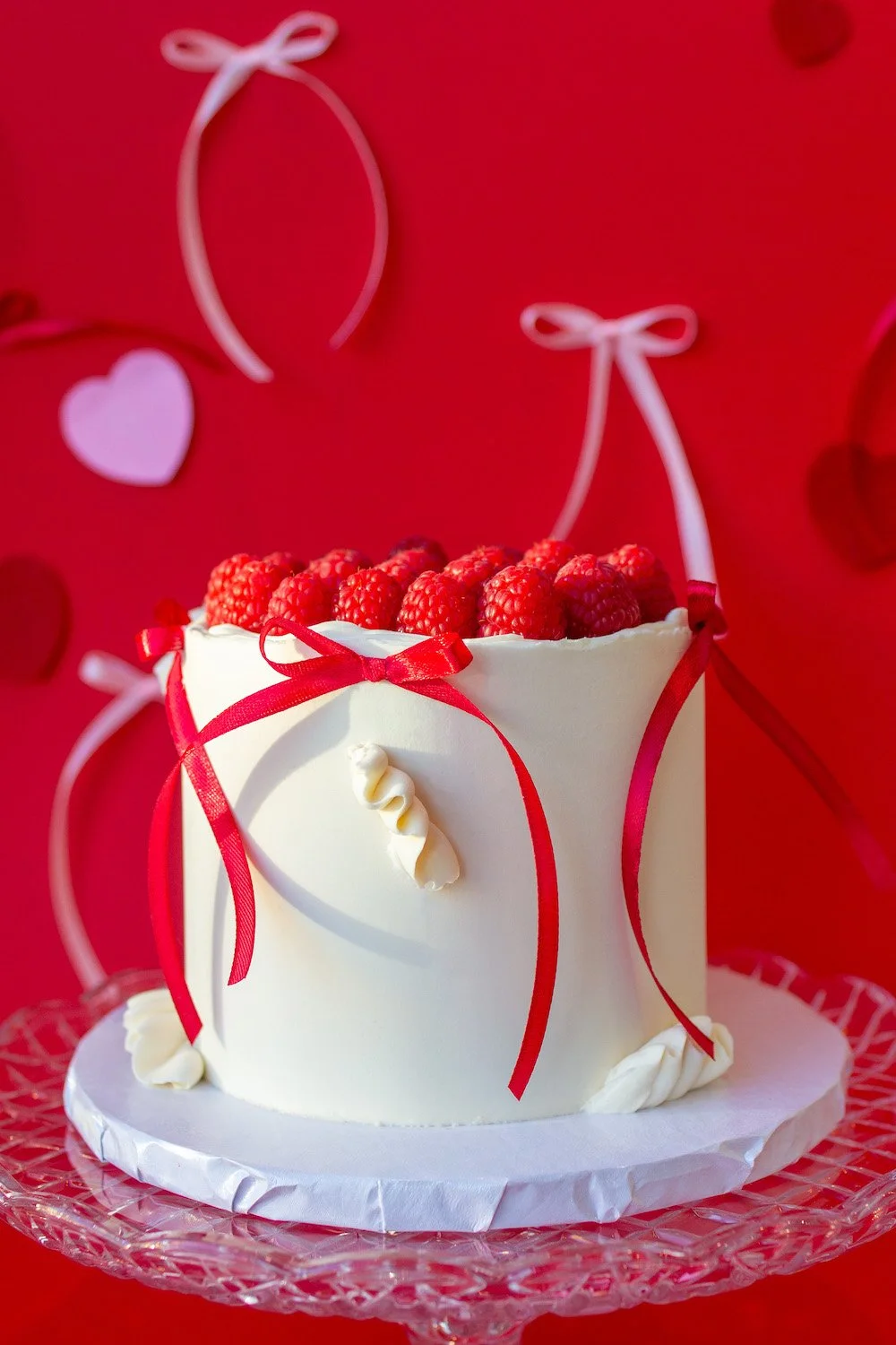 A white birthday cake decorated with raspberries on top, red ribbons, and creamy accents, positioned against a red background with heart and ribbon decorations.