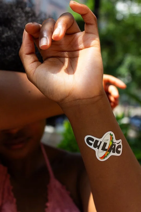 Close-up of a child's hand showing a temporary tattoo of the word "cubic" with a colorful stylized rainbow on the wrist. In the background, part of the child's face and upper body are visible, with green trees and outdoor lighting.