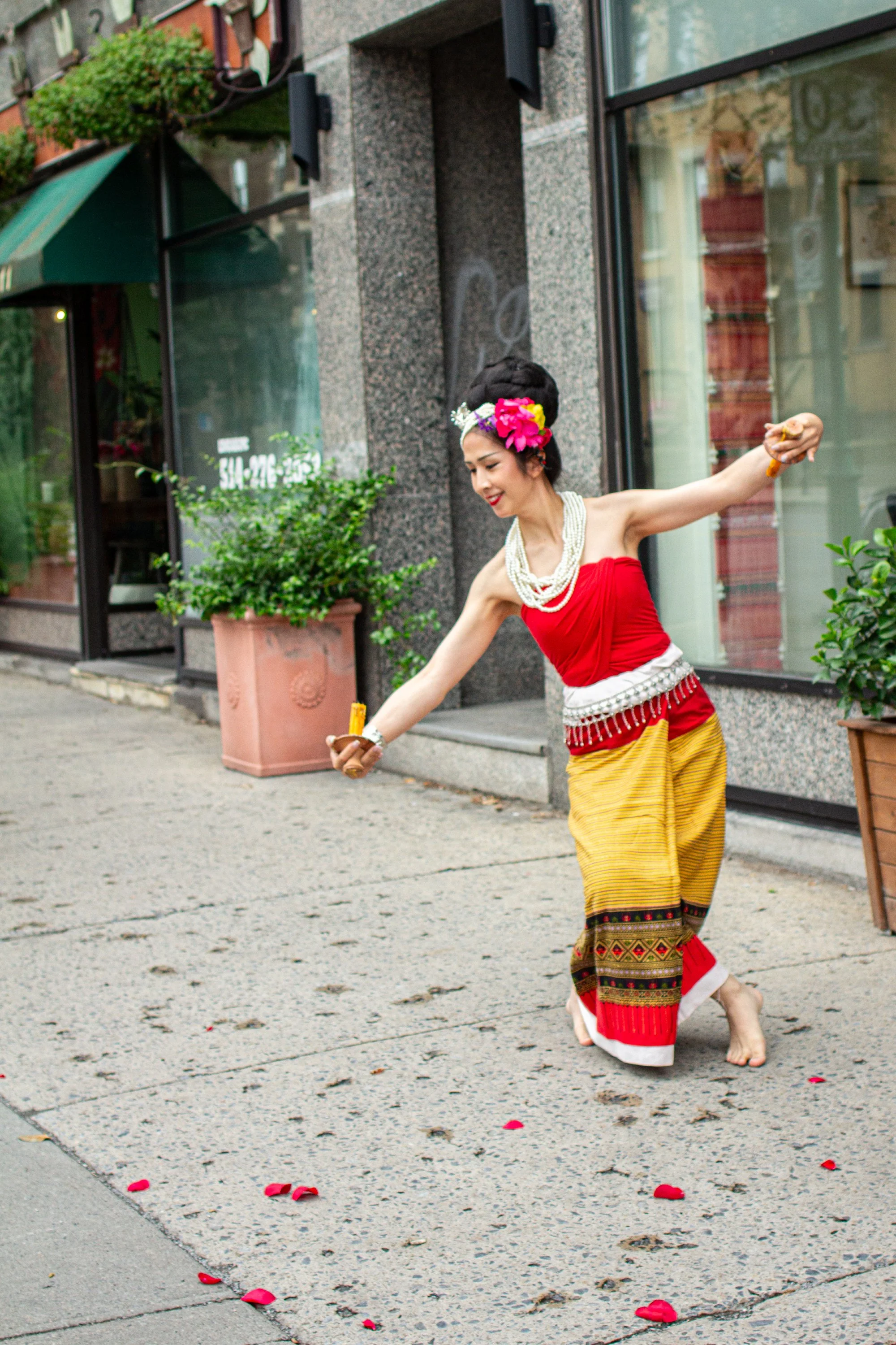 A woman performing a traditional dance on a city street, dressed in colorful cultural attire with flower accessories in her hair, surrounded by rose petals.