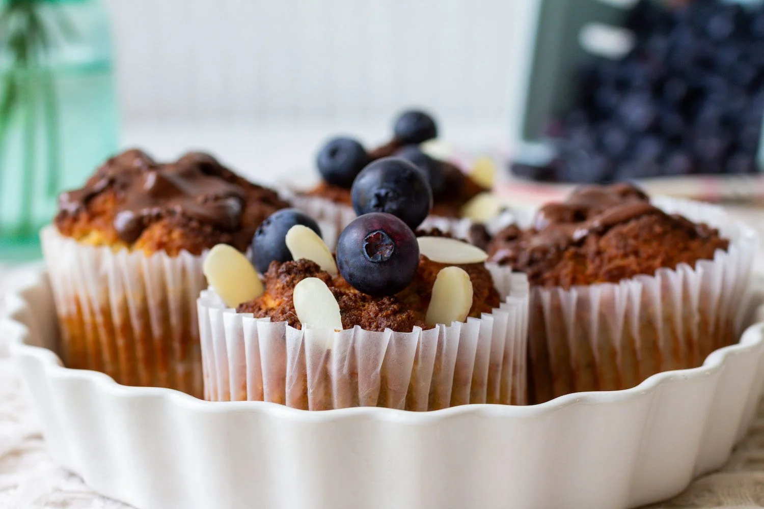 Cupcakes topped with blueberries and white almond slices in a white scalloped dish.