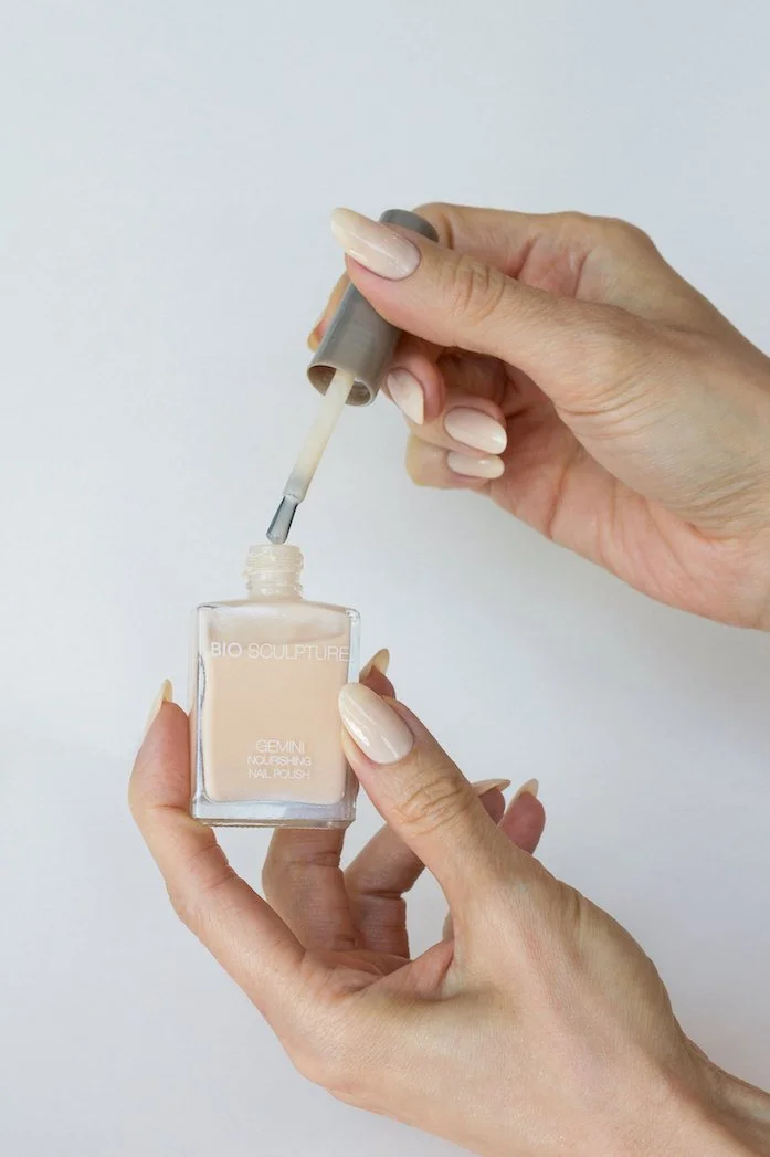 A person with manicured nails holding a bottle of nude-colored nail polish and applying it with a brush.