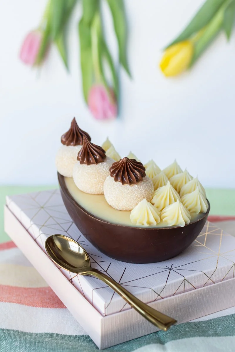 Chocolate and vanilla dessert served in a half coconut shell, garnished with chocolate and sugar-coated balls, whipped cream, and pink and yellow tulips in the background.