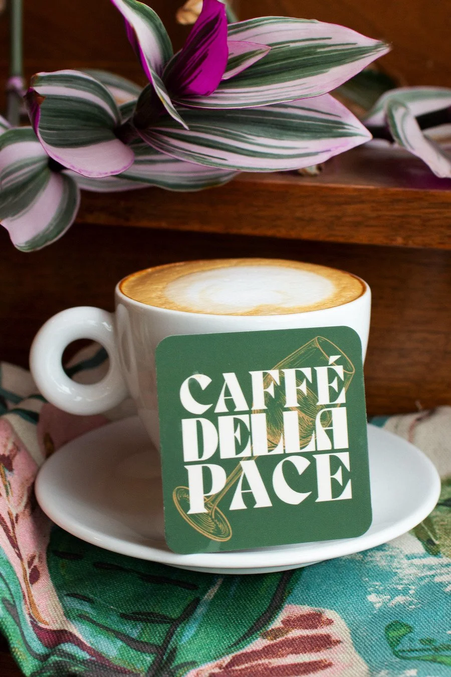 A white coffee cup with a latte that has foam art, placed on a matching saucer. The cup has a green and white sticker that says 'Caffé Della Pace'. The background includes a purple and white striped plant and a colorful fabric with a floral pattern.