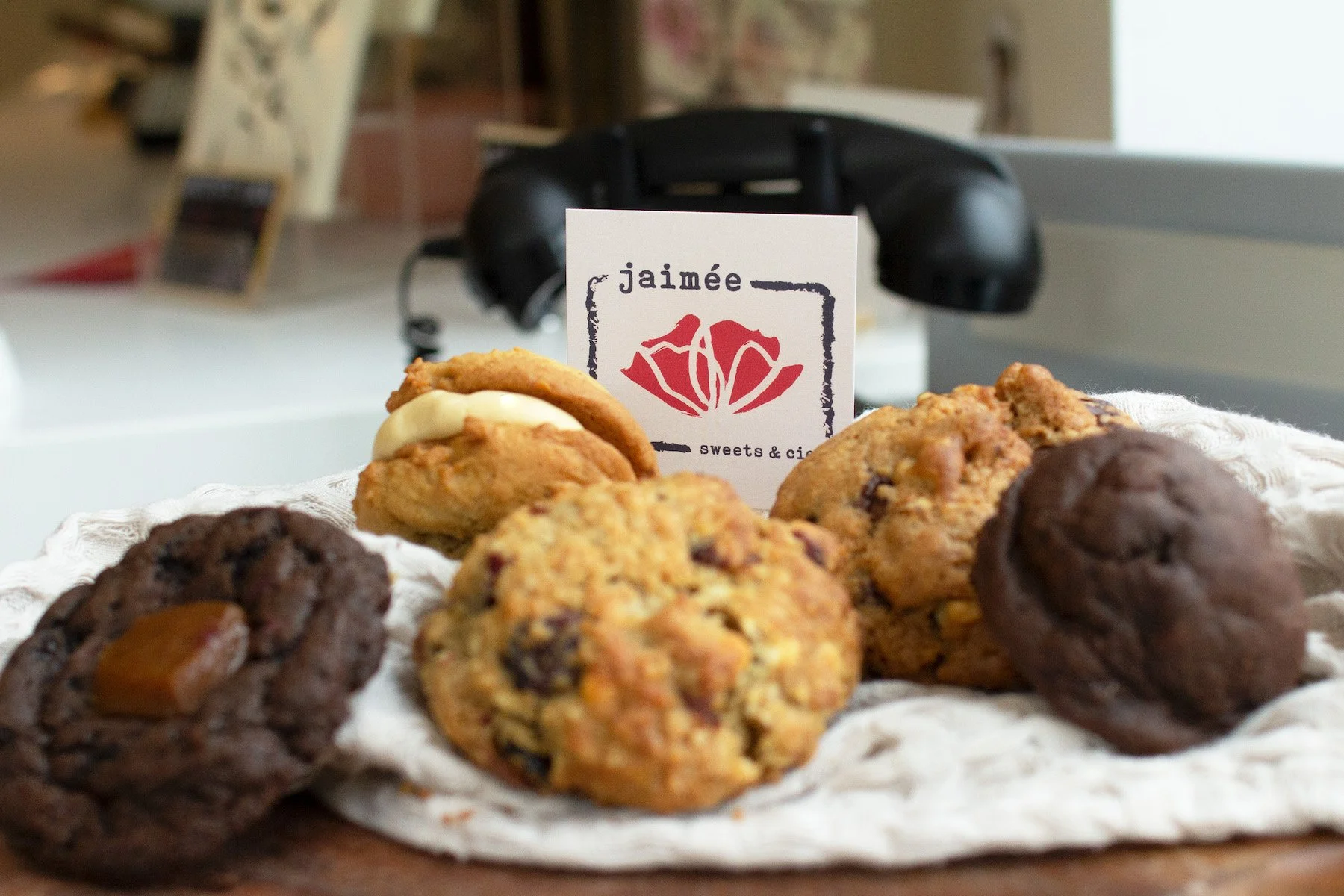 Assorted cookies on a white cloth, with a sign that reads 'jaimée sweets & cic', in front of a vintage black telephone.