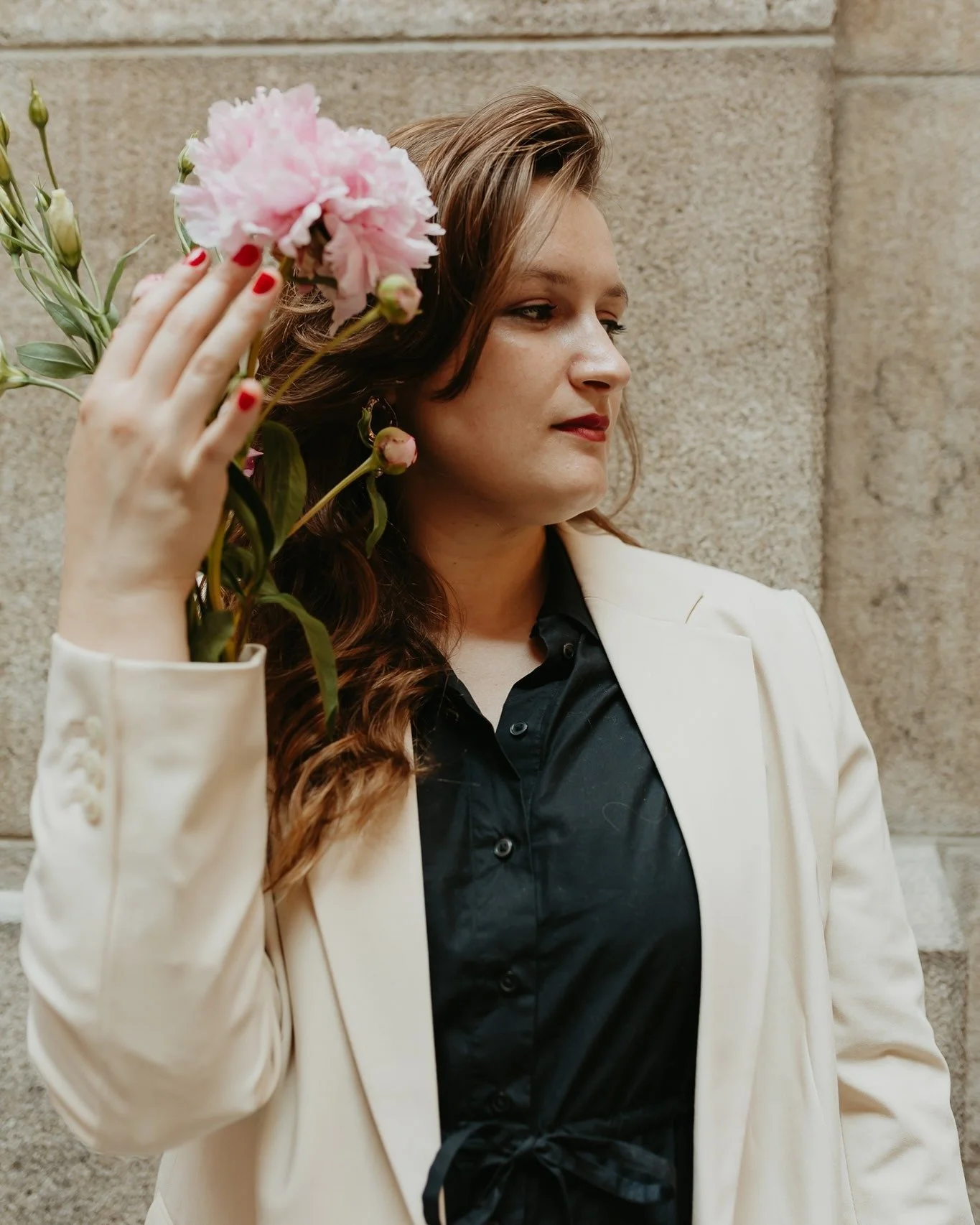 A woman with auburn hair in a white blazer and black blouse holding pink flowers near her face, standing against a brick wall.