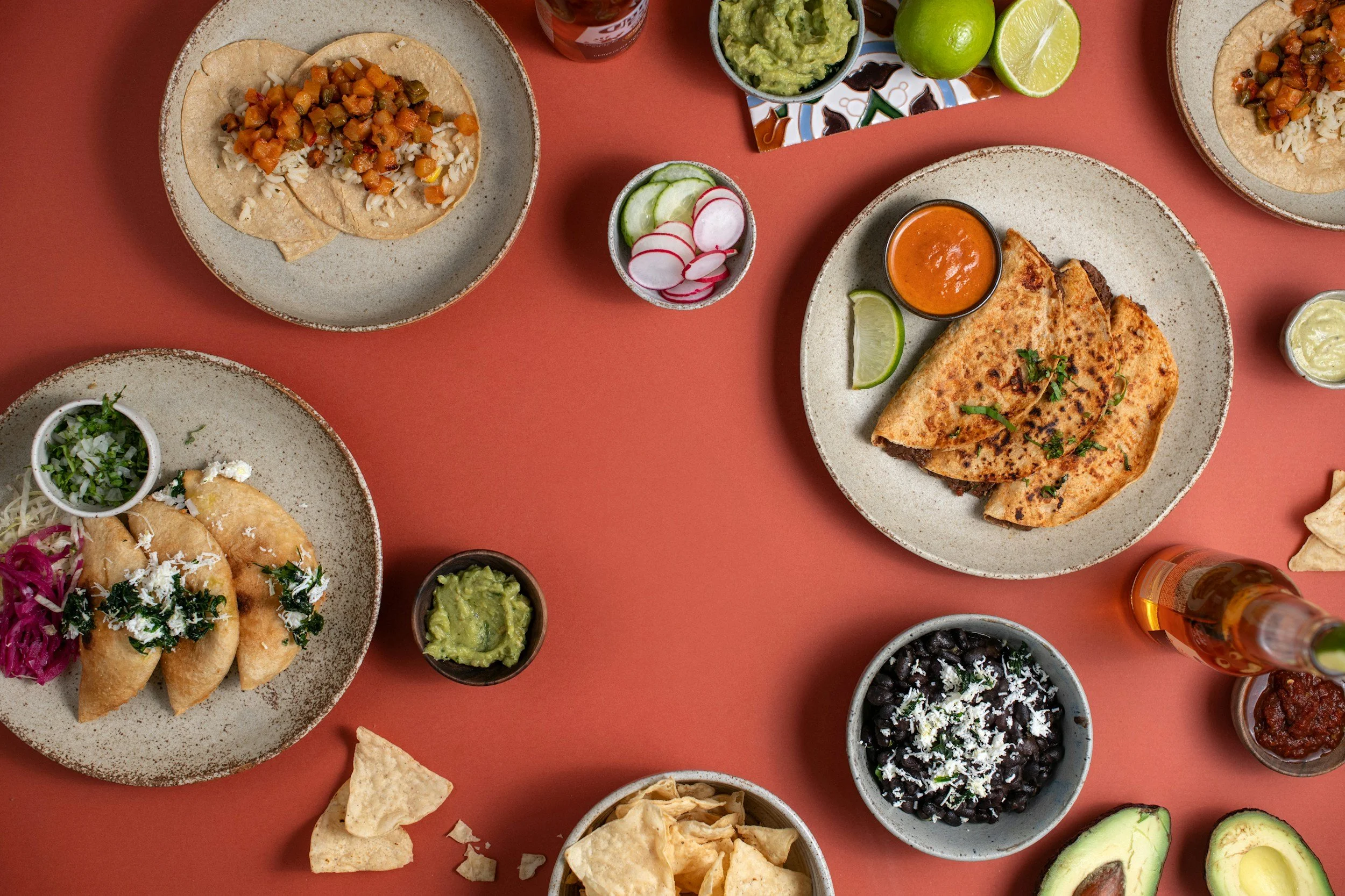 Assorted Mexican dishes on a reddish table, including tacos, quesadillas, and nachos, with guacamole, salsa, lime, radish, cucumber, and a beer.