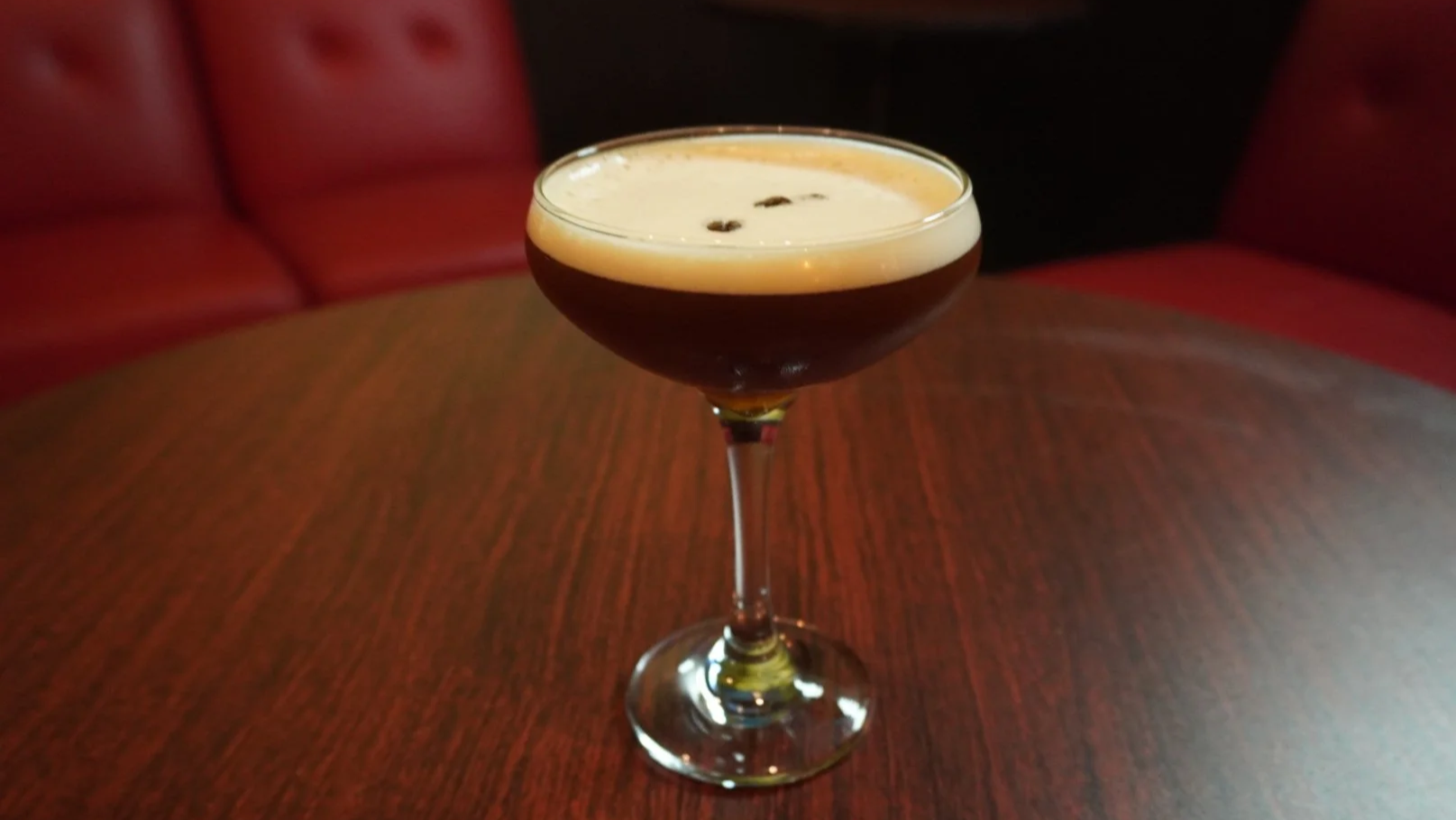 A dark cocktail with a foamy top and a few small dark ingredients in a coupe glass on a wooden table, with red upholstered seating in the background.