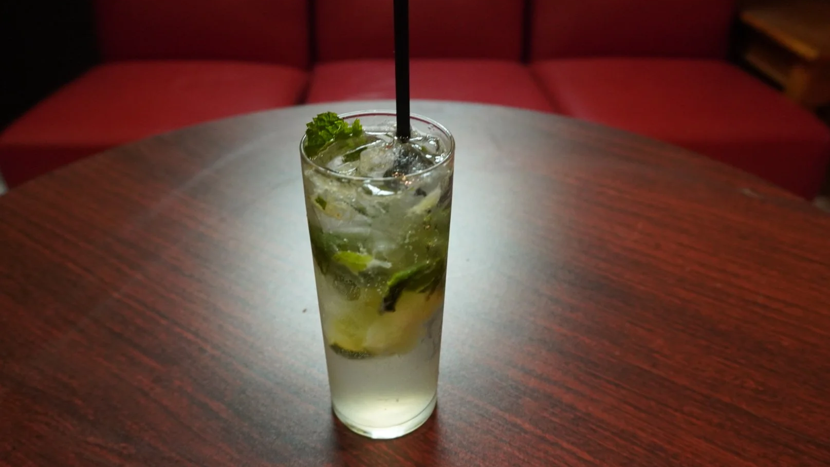 A tall glass of mojito cocktail with lime slices, mint leaves, ice cubes, and a black straw, on a wooden table in a dimly lit setting.
