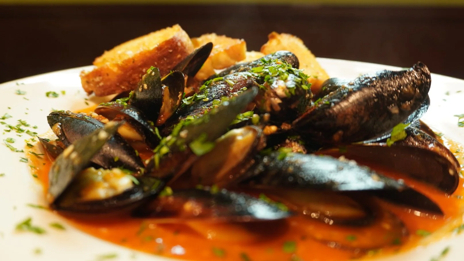 A plate of cooked mussels garnished with chopped herbs, served with slices of bread in the background.