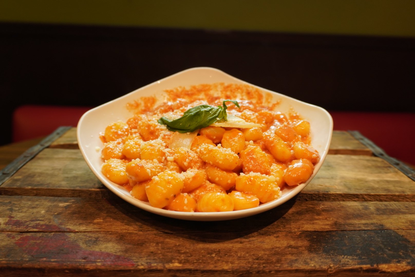 A white bowl of gnocchi with tomato sauce, topped with grated cheese and garnished with basil leaves, on a rustic wooden table.