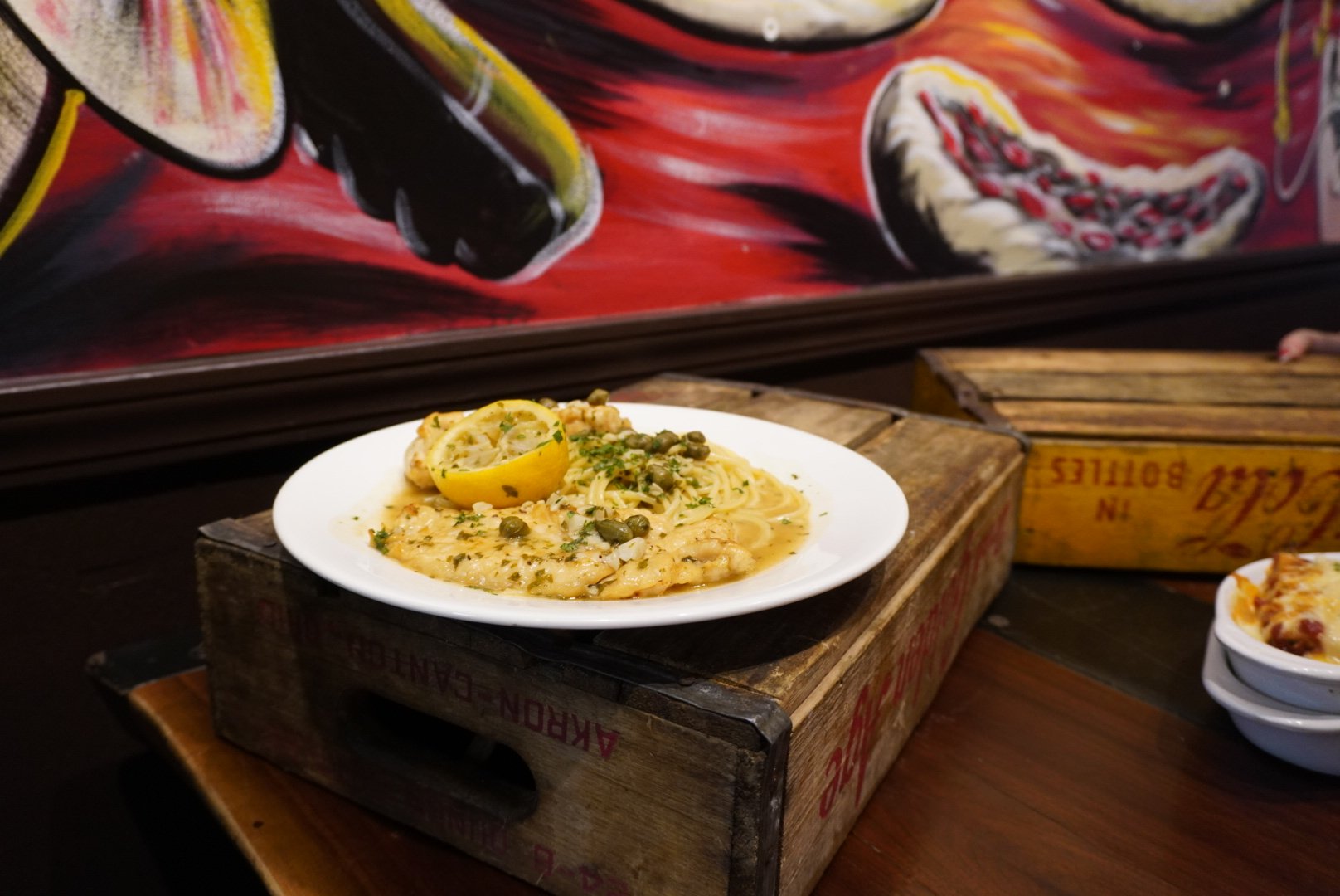 A white plate of creamy pasta garnished with herbs and served with a lemon wedge, placed on a wooden crate on a dark wooden table, with a colorful mural in the background.