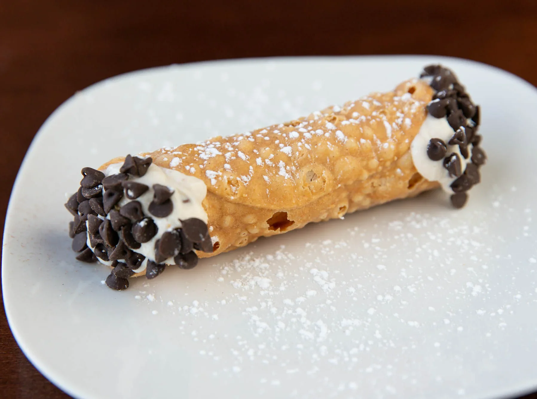 Cannoli with chocolate chips on the ends and powdered sugar dusted on top, served on a white plate.