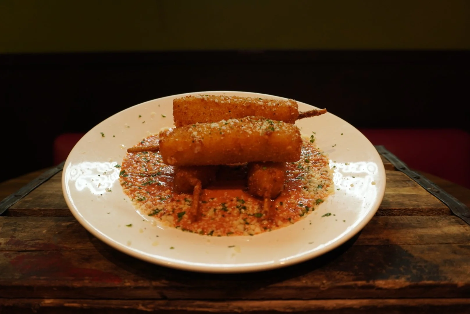 Plate of fried mozzarella sticks served on marinara sauce, garnished with grated cheese and herbs, on a white plate placed on a wooden surface.