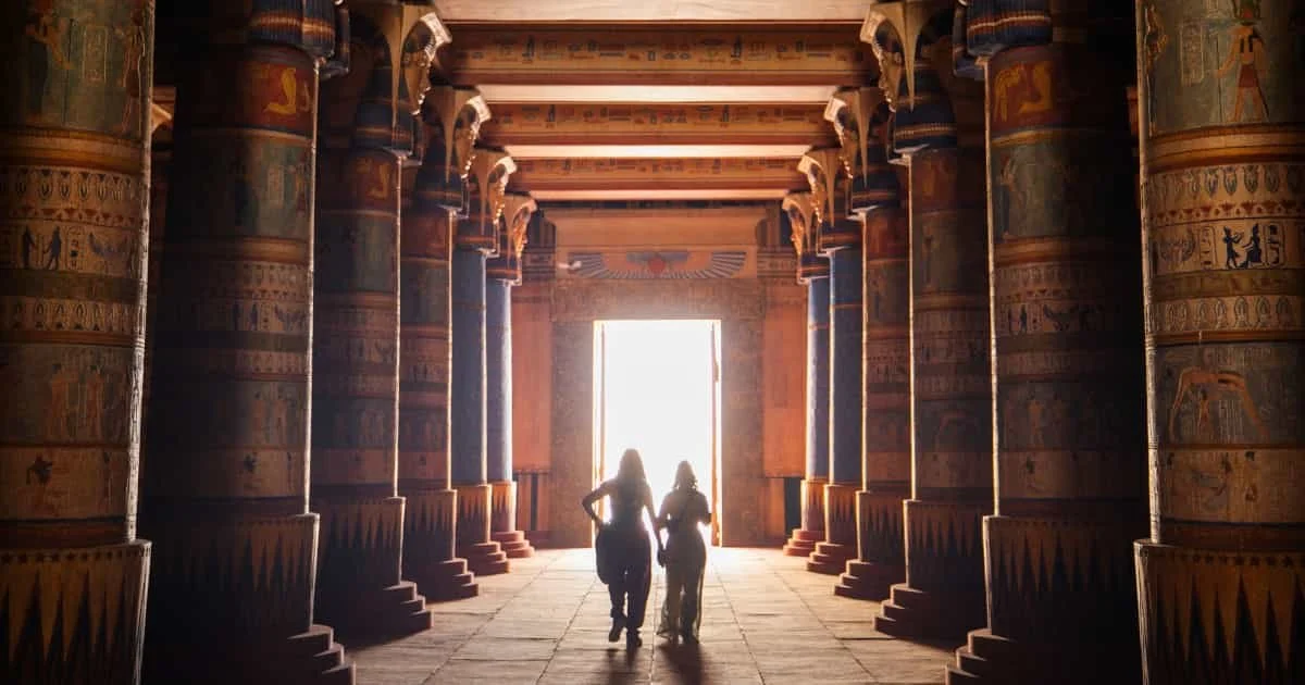 Vista cinematográfica del interior de un palacio egipcio con columnas pintadas, jeroglíficos y dos siluetas femeninas avanzando hacia la luz