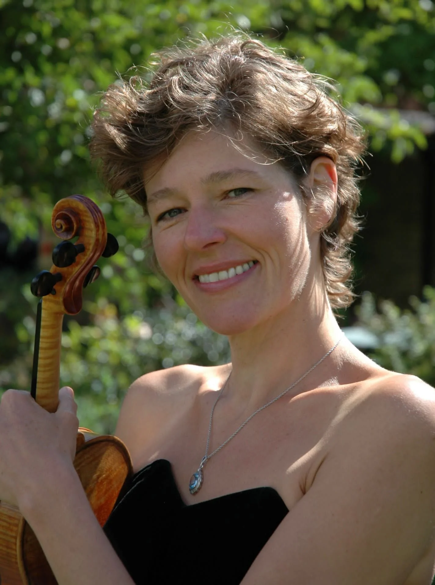 A woman with short curly hair smiling and holding a violin outdoors with green foliage in the background.
