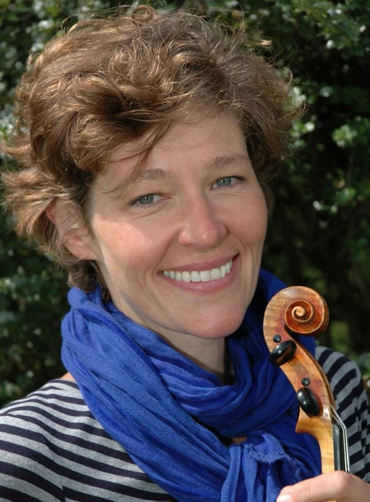 A woman with short curly red hair smiling and holding a violin, outdoors with greenery in the background.